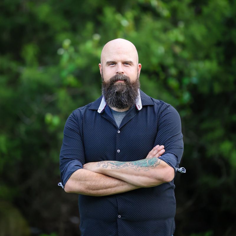 Bearded bassist standing arms crossed in front of trees